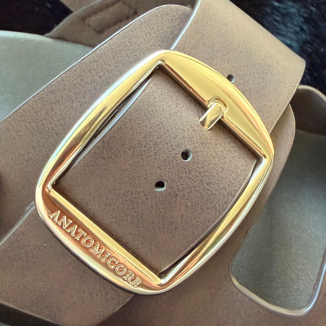 A close-up of a taupe brown leather sandal with a large, polished gold-tone metal buckle engraved with the brand name 'ANATOMICORE'.