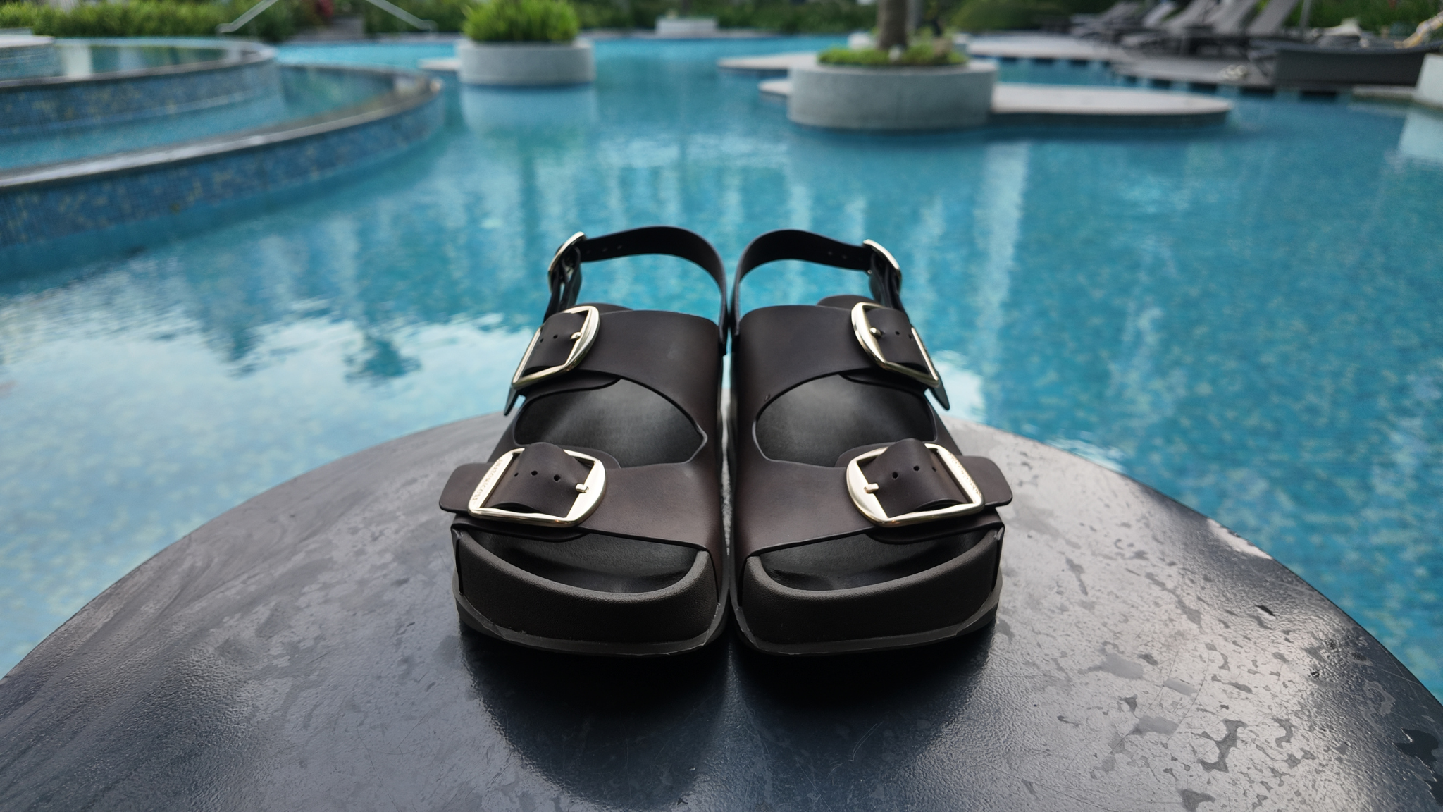 Brown sandals with gold buckles on a stone ledge by a pool.